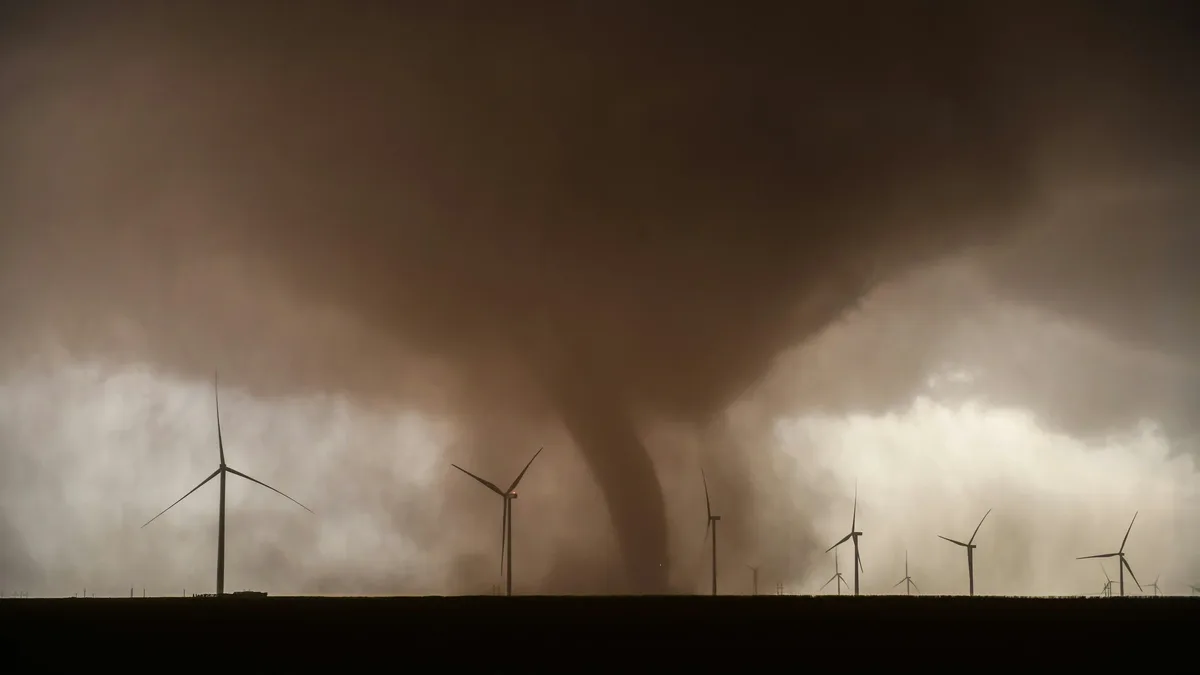 A tornado descending from a dark, stormy sky, with wind turbines visible in the foreground.